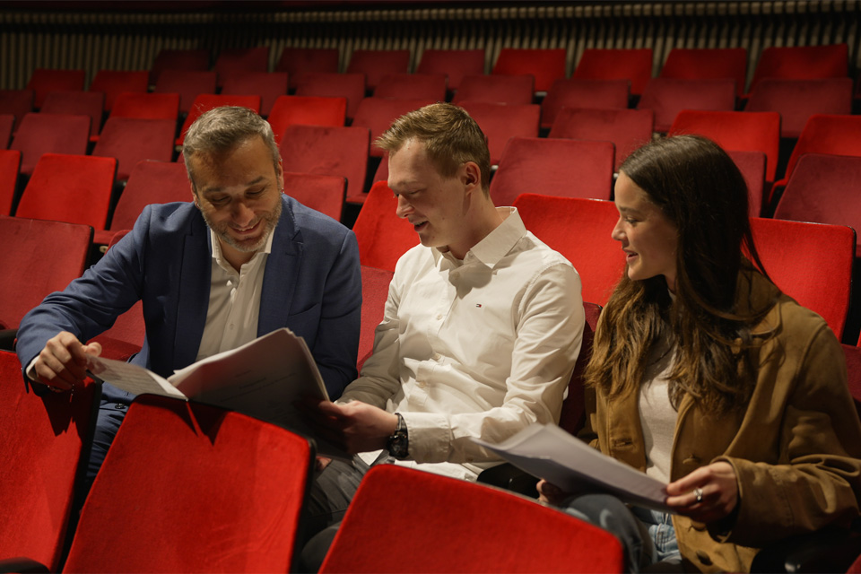 A man wearing a suit talking to a male and female students, looking at their music sheets, sitting in red theatre chairs.